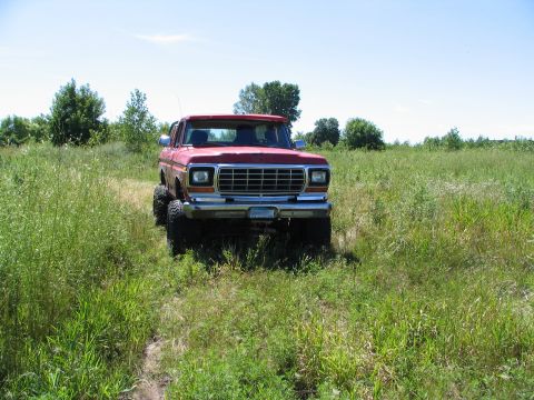 1978 Ford Bronco - A red 1978 Ford Bronco is shown from the front, lifted and driving through a grassy field under a clear blue sky.