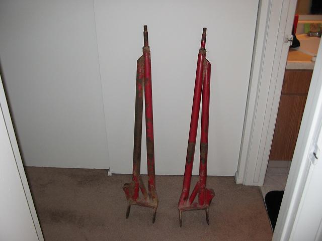 Two red metal jack stands with rusted sections, standing on a carpeted floor against a white wall
