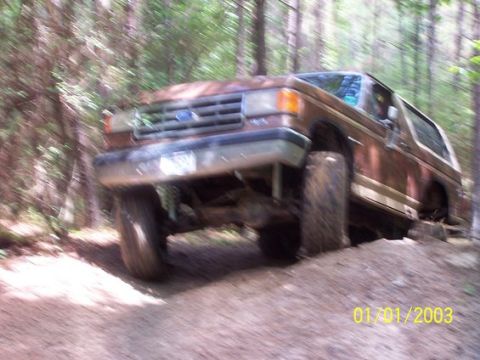 1987 Ford Bronco - A brown two-tone full-size Ford Bronco flexes over a dirt mound on a wooded trail, front driver's wheel drooping and the body leaning as it…