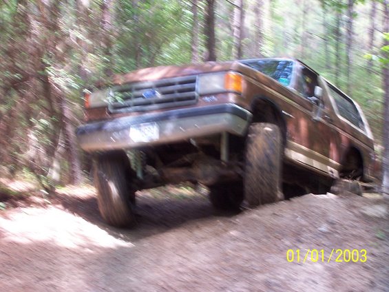 Brown lifted Ford Bronco flexing over a dirt mound on a wooded trail