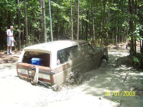 1987 Ford Bronco - Mud-covered Ford Bronco buried to its rocker panels in a wooded mud hole, shot from the rear three-quarter with the rear glass removed and…