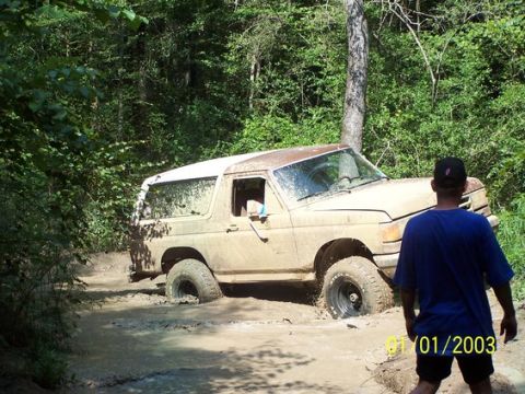 1987 Ford Bronco - A tan 1987 Ford Bronco is stuck deep in mud on a wooded trail, with a man observing from the side.