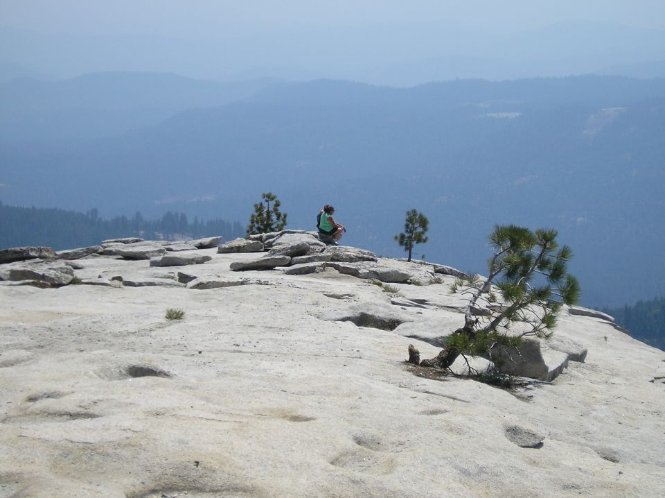 Person sits on a rocky summit with trees, overlooking a hazy blue valley