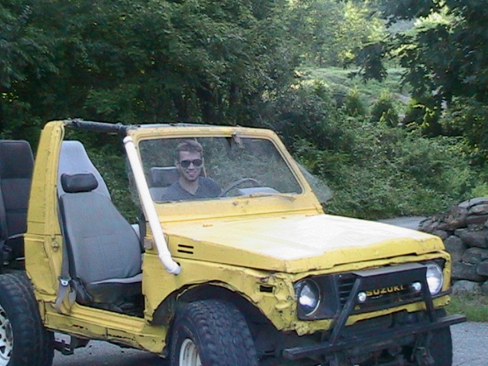 Yellow 1987 Suzuki Samurai with a man driving. It has a roll bar and off-road tires