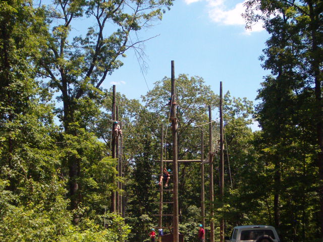Ropes course with people climbing wooden poles in a wooded area