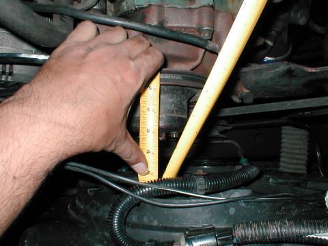 1979 Ford Bronco - Close-up of a hand measuring an engine part with a yellow ruler, with hoses and wiring visible.