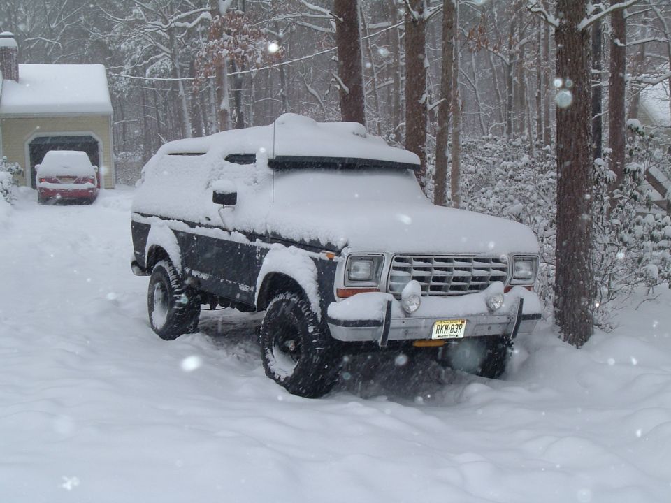 Dark blue Ford Bronco covered in snow, parked in a snowy driveway with a red car in the background garage