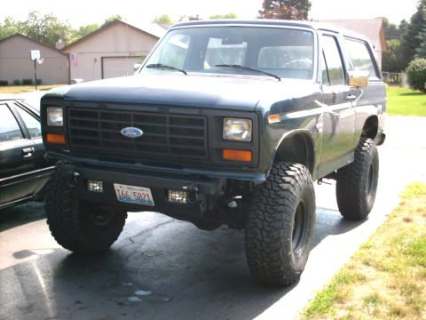 1986 Ford Bronco - A dark green/blue 1980s Ford Bronco full-size SUV lifted on large mud-terrain tires, shown from a three-quarter front angle in a driveway.…