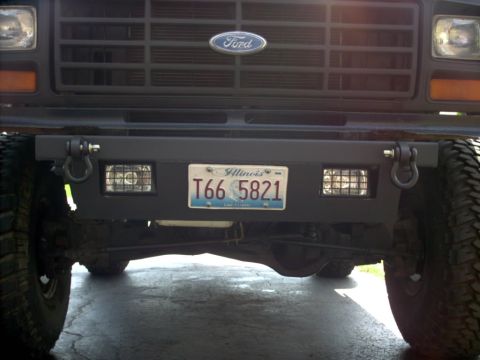 1986 Ford Bronco - Front view of a Ford truck's lower front end, showing a gray steel bumper with D-ring shackles, dual auxiliary grille-guarded lights, and a…