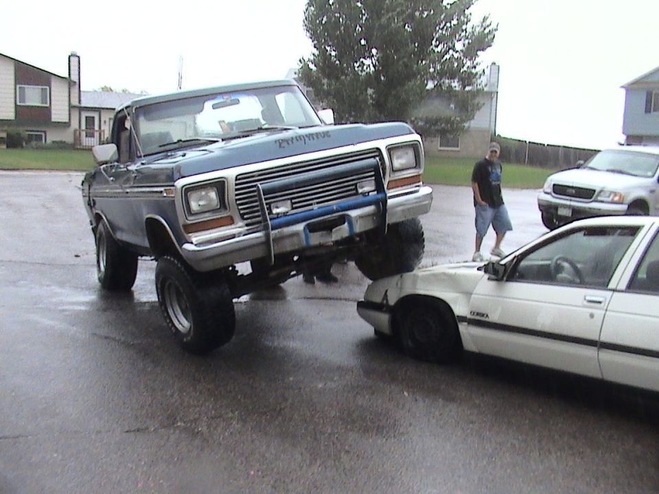 Blue 1979 Ford Bronco with a brush guard on top of a crushed white sedan