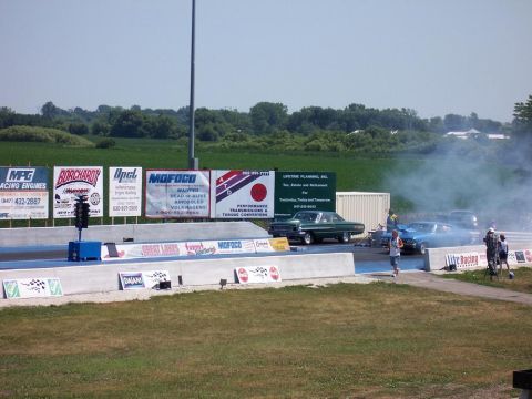1964 Ford Galaxie - Two cars prepare for a drag race on a sunny day, with advertising banners in the background.