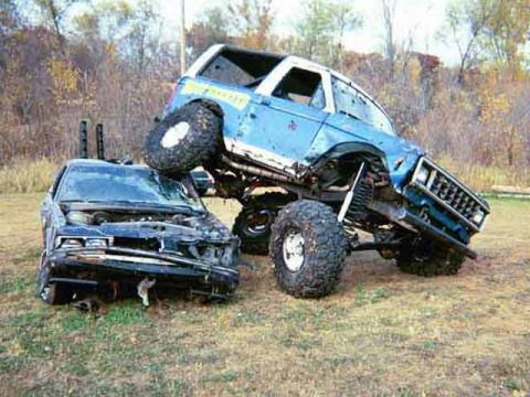 1985 Ford Bronco - A blue 1985 Ford Bronco is perched atop a severely damaged car, a dramatic scene.
