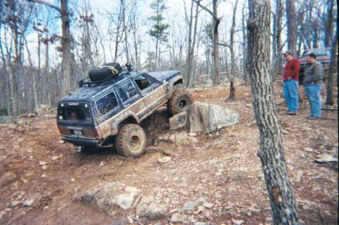 1985 Ford Bronco - A modified Jeep Cherokee XJ navigates a rocky obstacle on an off-road trail, with two onlookers in the background.