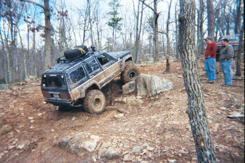 Black Jeep Cherokee XJ with roof rack and spare tire climbing over rocks on a trail