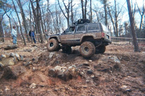 1985 Ford Bronco - Jeep Cherokee XJ navigating a challenging off-road course in a wooded area.