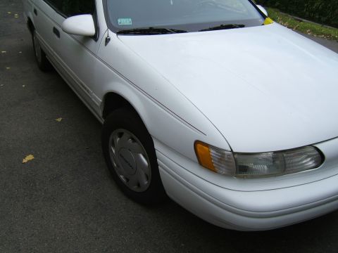 1998 Ford Taurus - The front quarter panel and headlight assembly of a white sedan are visible, parked on an asphalt surface.