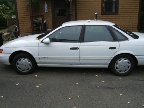 1998 Ford Taurus - Side view of a white sedan with a can of spray on its roof, parked on a paved surface with scattered leaves.