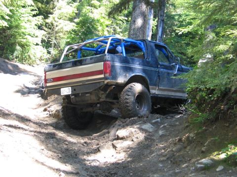 1989 Dodge Power Ram - A dark blue truck with a roll cage navigates a challenging dirt trail in a wooded area.