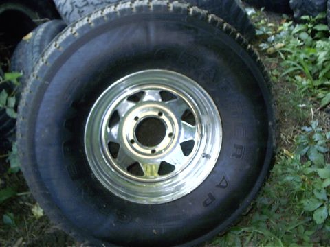 1979 Ford Bronco - A spare tire with a chrome rim is shown, with other tires and greenery in the background.