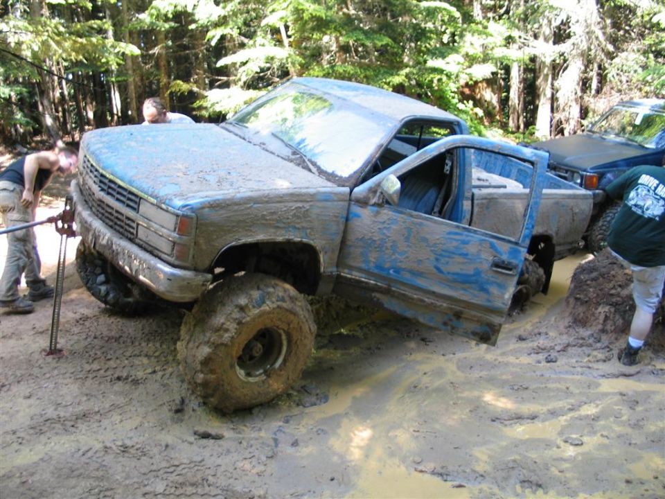 Lifted blue pickup on big mud tires stuck in a muddy forest trail with people helping.