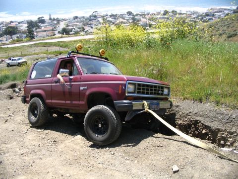 1985 Ford Bronco II - Maroon 1985 Ford Bronco II is stuck off-road, with a white pickup truck visible in the distance.