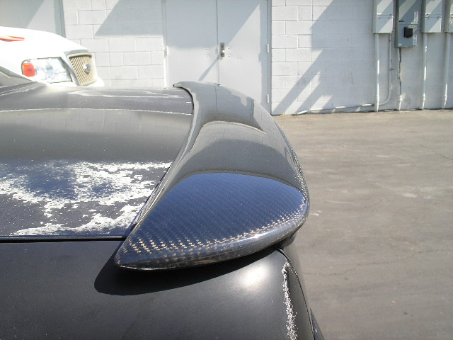 Carbon fiber rear spoiler mounted on a car's trunk lid, showing peeling paint on the trunk