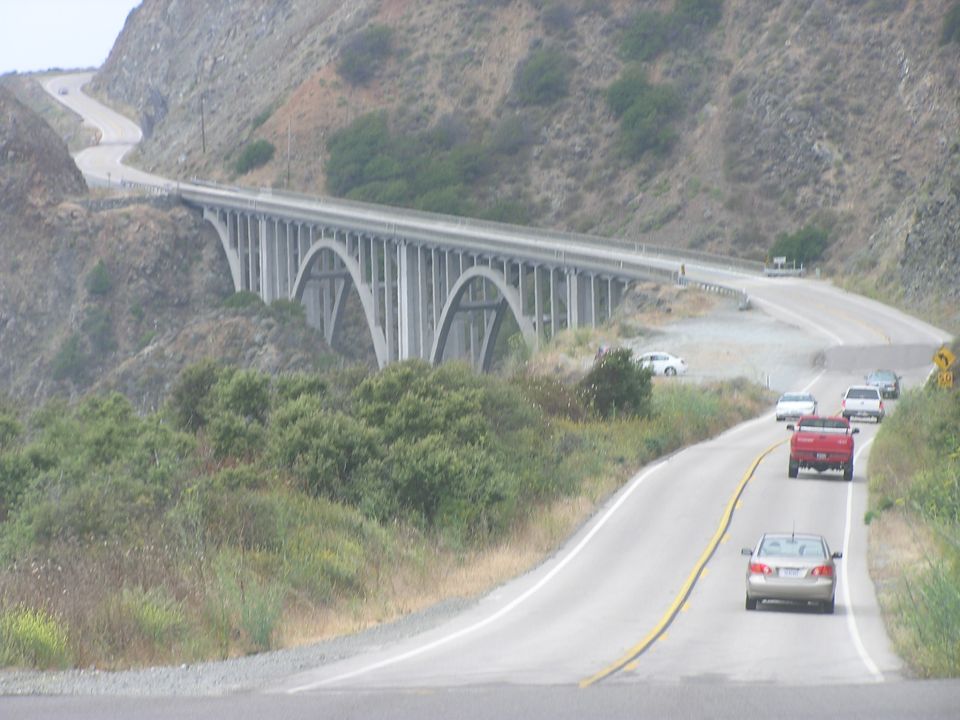 Several cars drive on a winding road near a large concrete arch bridge
