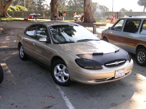 1993 Ford Taurus - A gold sedan is parked next to a wood-paneled van at a park gathering.