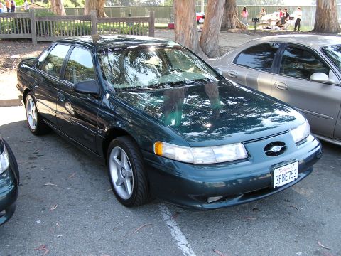 1993 Ford Taurus - This dark green sedan, likely a 1996 model, is parked next to another vehicle in an outdoor lot.