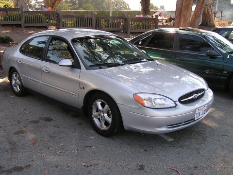 1993 Ford Taurus - A silver sedan is parked in an outdoor lot with a wooden fence and trees in the background.