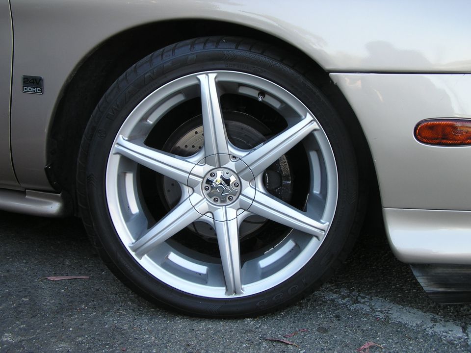 Close-up of a silver 5-spoke aftermarket wheel on a car, showing the tire, brake rotor, and part of the fender with a '24V DOHC' badge