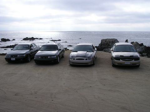 1993 Ford Taurus - Four Ford Taurus sedans are parked on a sandy area overlooking the ocean, with two dark and two lighter colored vehicles visible.