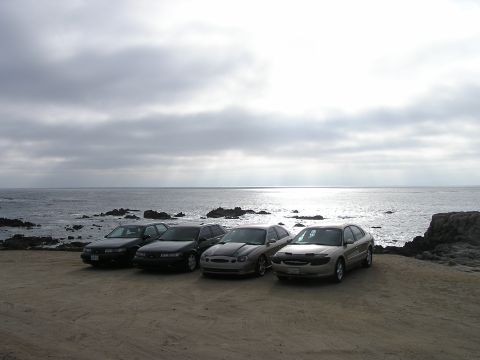 1993 Ford Taurus - Four sedans are parked on a dirt lot with a rocky coastline and ocean in the background under a cloudy sky.