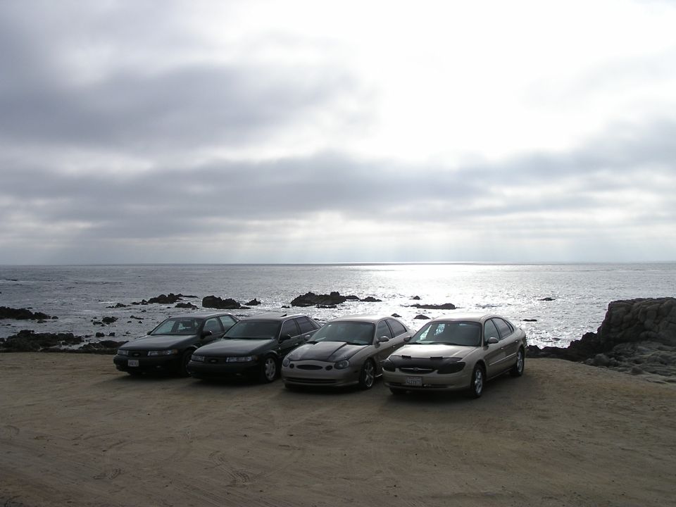 Four sedans parked on a dirt lot overlooking the ocean, one is a 2000 Ford Taurus