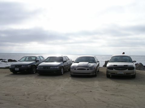 1993 Ford Taurus - Four sedans are parked on a beach with the ocean in the background under a cloudy sky.
