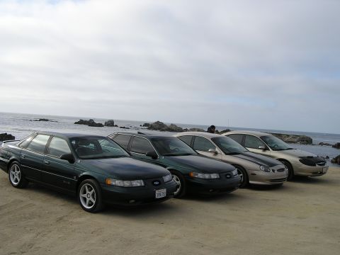 1993 Ford Taurus - A line of four Ford Taurus sedans are parked on a sandy area near the ocean.