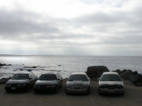 1993 Ford Taurus - Four sedans are parked in a line on a dirt lot overlooking the ocean under a cloudy sky.