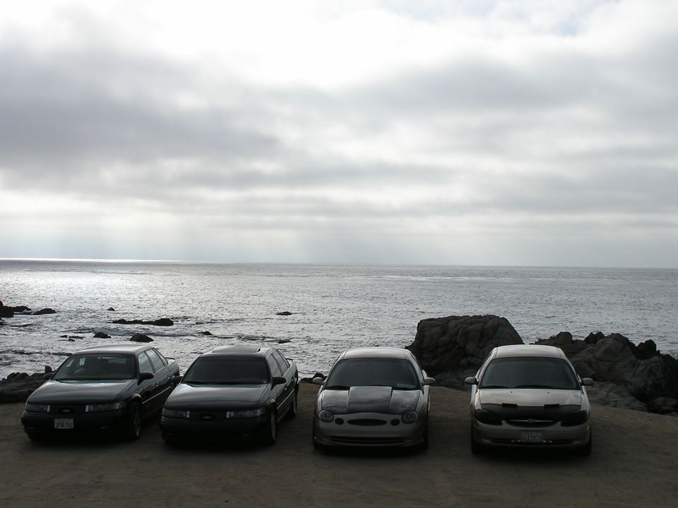 Four sedans parked on a dirt lot facing the ocean, two dark, one silver with a hood scoop, one tan with car covers