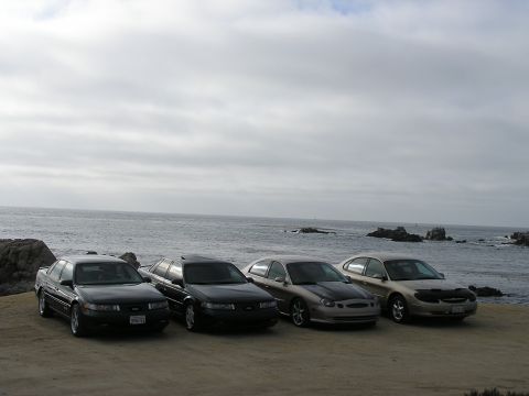 1993 Ford Taurus - Four sedans are parked on a dirt lot overlooking the ocean on a cloudy day.