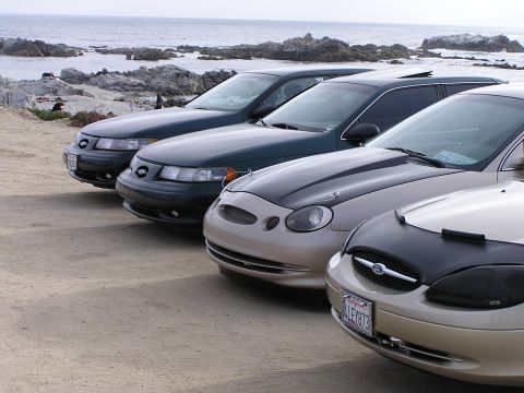 1993 Ford Taurus - Multiple Ford Taurus sedans are parked along a rocky coastline under an overcast sky.