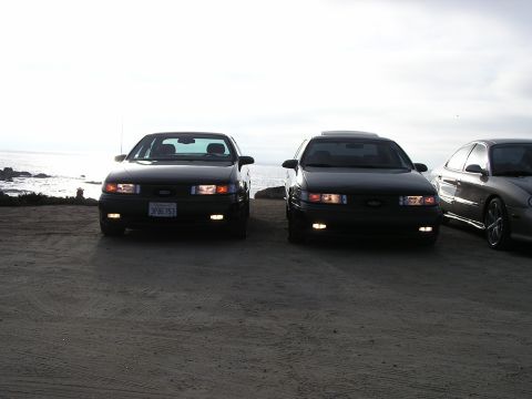 1993 Ford Taurus - Two dark-colored sedans are parked on a dirt lot facing the ocean.