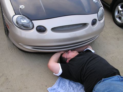 1993 Ford Taurus - A man lies on a towel beneath the front of a tan car with a black hood, while another vehicle is partially visible.