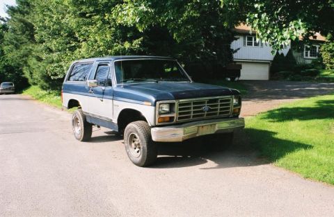 1978 Ford Bronco - A two-tone blue and silver Ford Bronco is parked on a street in front of a house and trees.