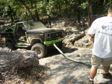 1995 Ford Bronco - A black off-road vehicle with a green roll cage and lime green bumper is being pulled by a strap through a rocky, wooded area.