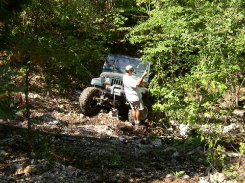 1995 Ford Bronco - A person stands next to a green Jeep Wrangler on a rough, wooded terrain.