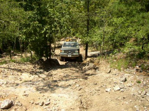 1995 Ford Bronco - An SUV navigates a rough, unpaved path surrounded by trees, with another vehicle in the background.