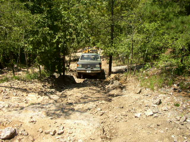 SUV driving on a rocky, wooded trail. Another vehicle is visible behind it