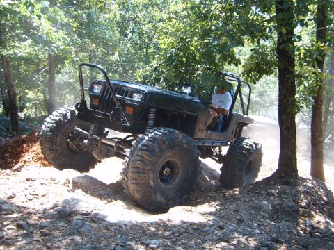 1988 Ford Bronco - A heavily modified black Jeep Wrangler navigates a rocky, wooded trail with its large tires.