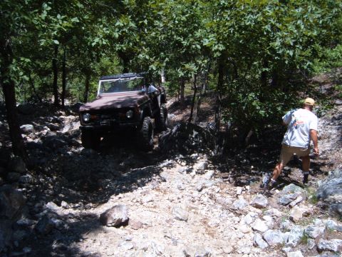 1988 Ford Bronco - A person watches a classic Ford Bronco navigate a challenging, rocky off-road path surrounded by dense foliage.