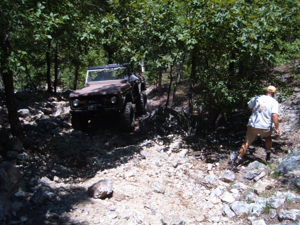 Brown Ford Bronco driving on rocky trail with trees around. Person walking behind it